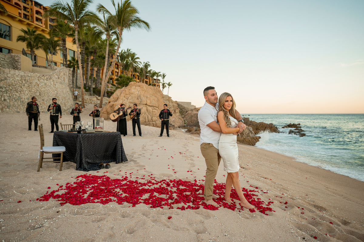 A couple embraces on a beach scattered with red rose petals, mariachi band and hotel behind them at Fiesta Americana Los Cabos