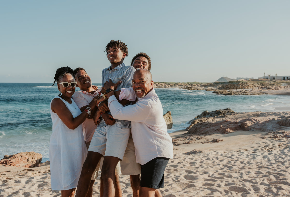 Family of five embracing and laughing on a rocky beach at Fiesta Americana Los Cabos, ocean behind them