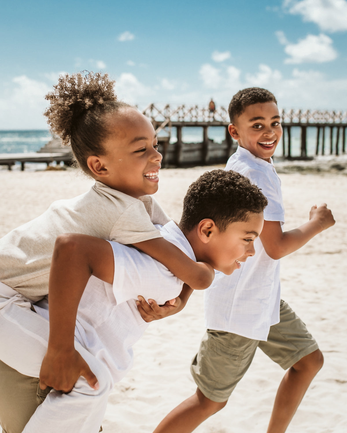 Three laughing kids in white shirts play piggyback on the beach near a wooden pier during a family photoshoot