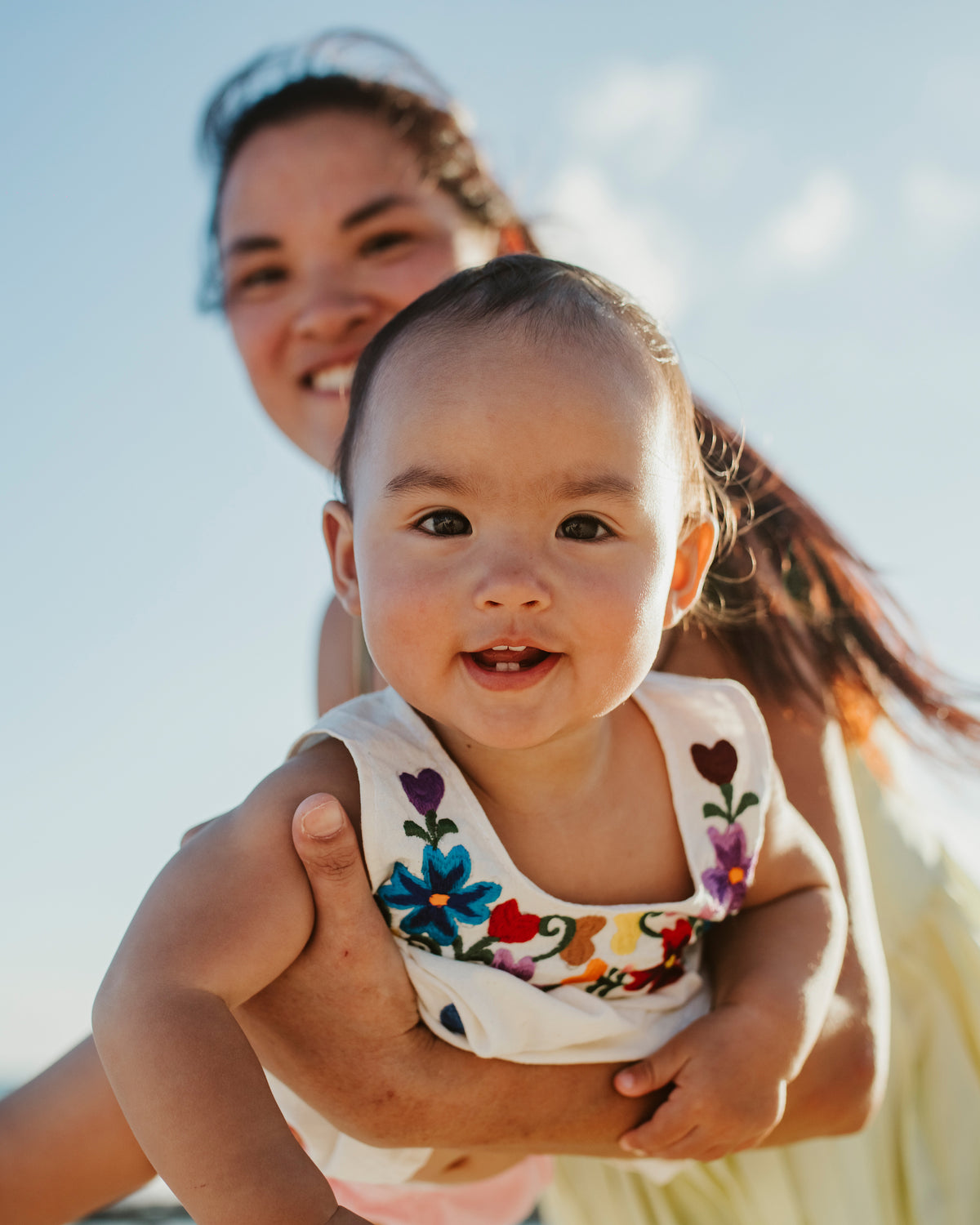 Smiling baby in an embroidered dress held up by her mother under a blue sky during a family photoshoot