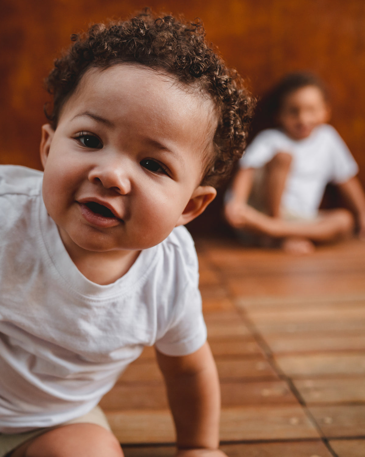 Curly-haired toddler crawls toward the camera on a wooden deck while a sibling sits behind during a family photoshoot