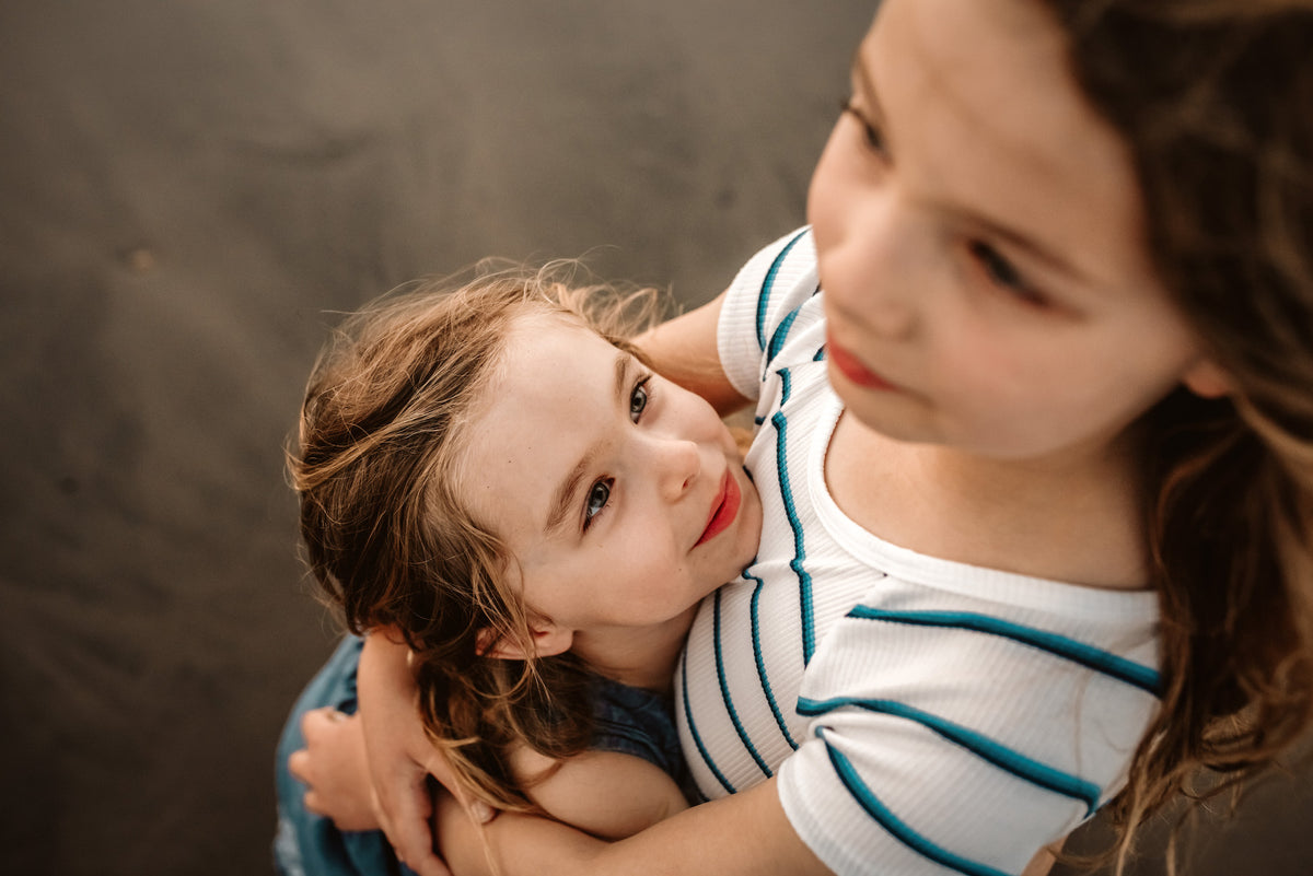 Little girl gazes up smiling as her big sister hugs her in an overhead shot on the sand