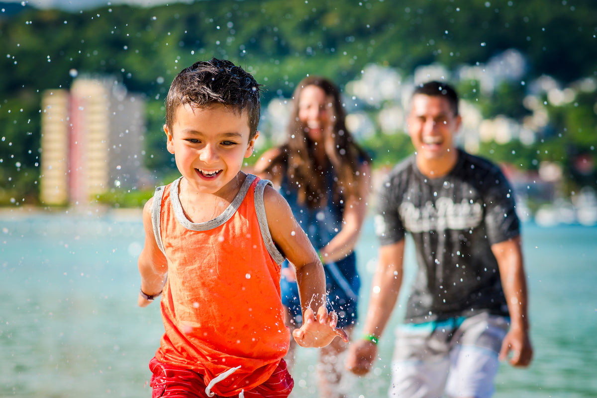 A boy in an orange tank top runs through splashing water with adults smiling behind him