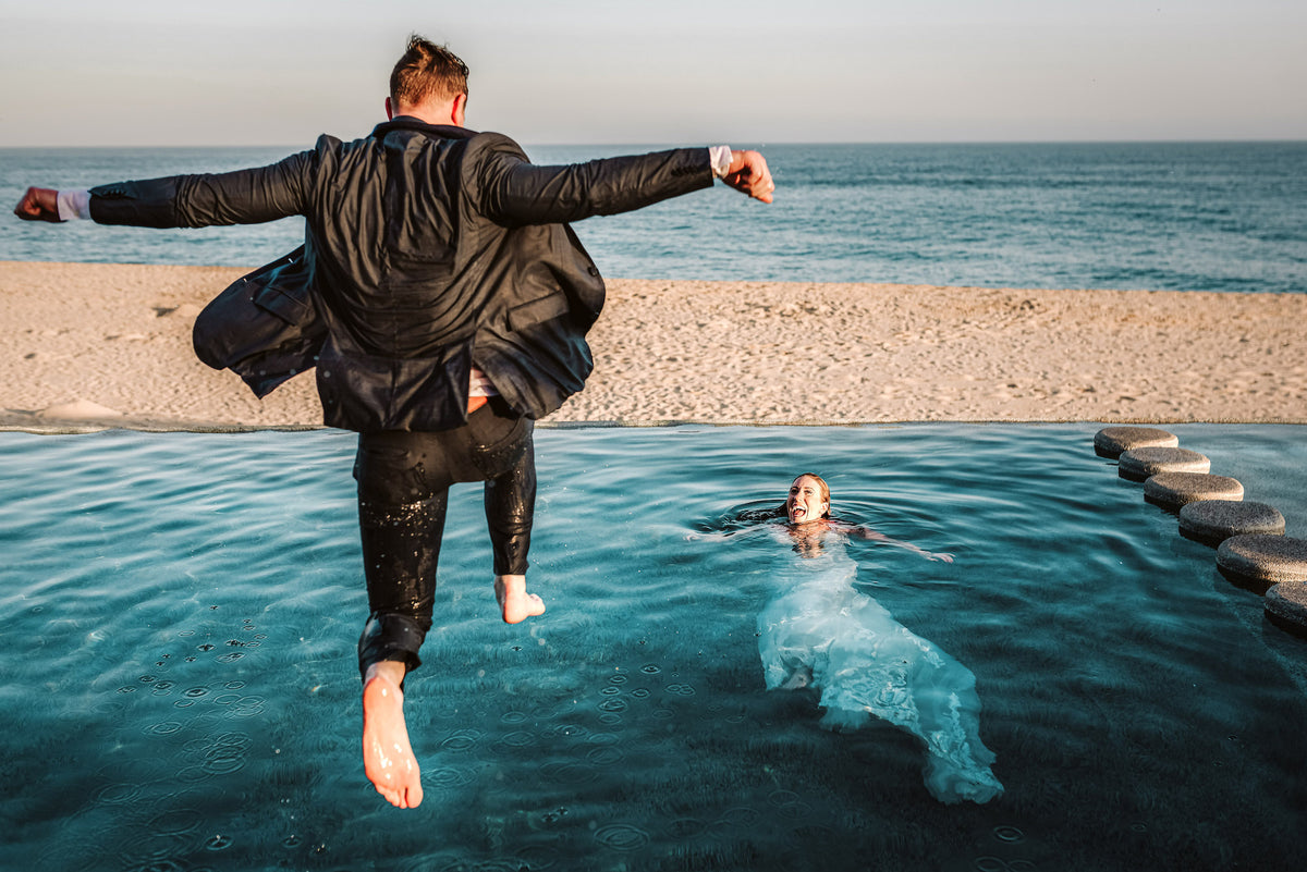 A man in a suit jumps into a pool as a woman in a wedding dress swims nearby at Marquis Los Cabos, ocean visible beyond the pool edge