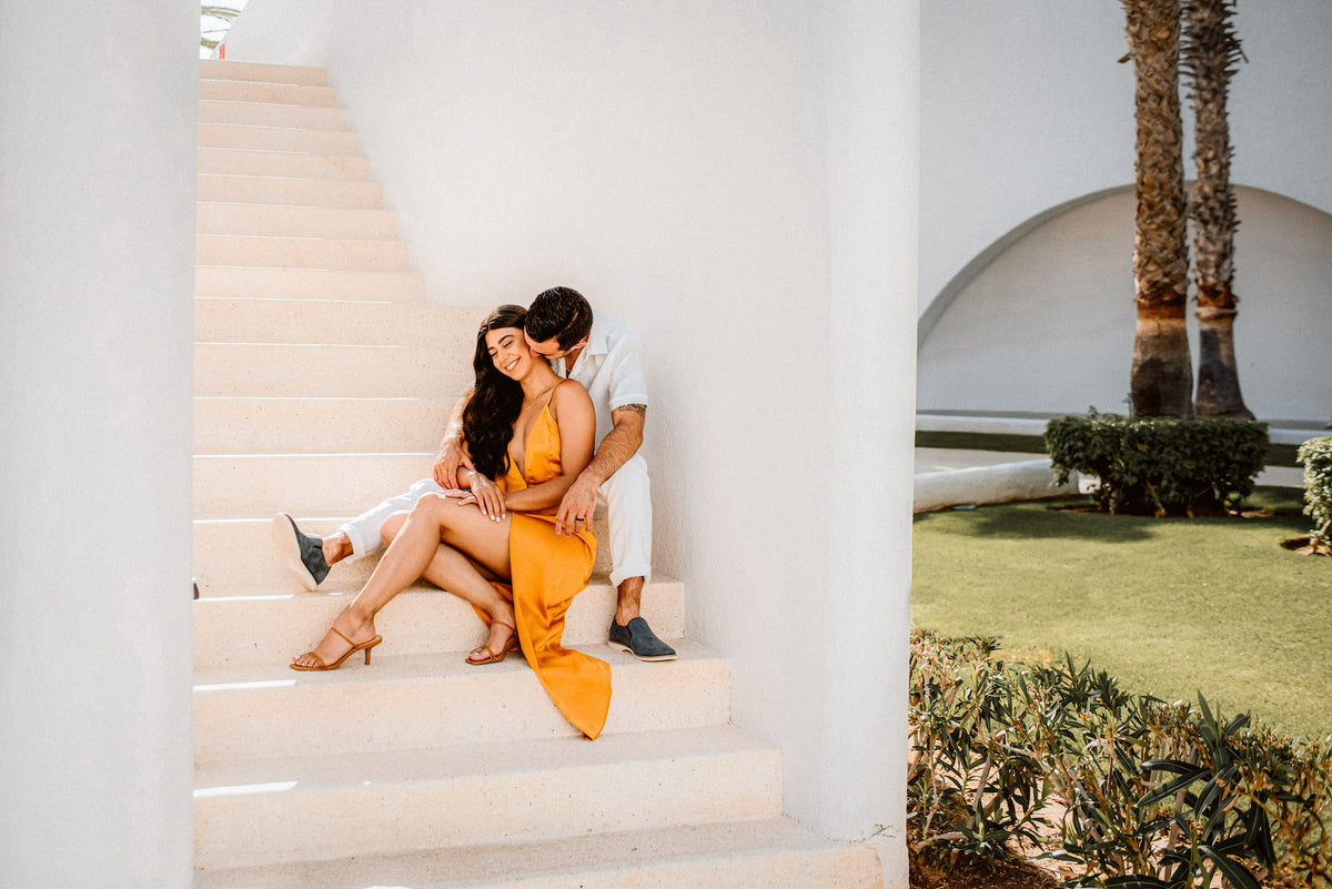 A couple embraces on white steps at Marquis Los Cabos, palm trees visible beyond the wall