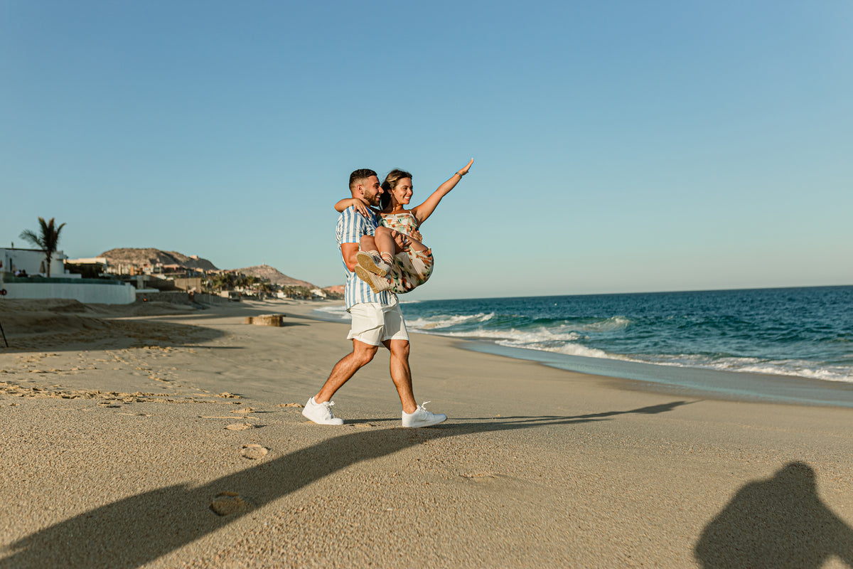 A man carries a woman on a beach at Marquis Los Cabos as she reaches toward the ocean