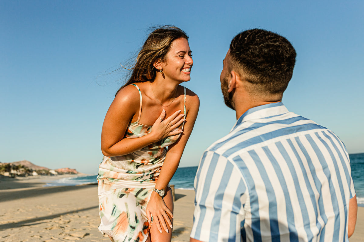 A woman laughs with her hand on her chest while a man faces her on a Los Cabos beach