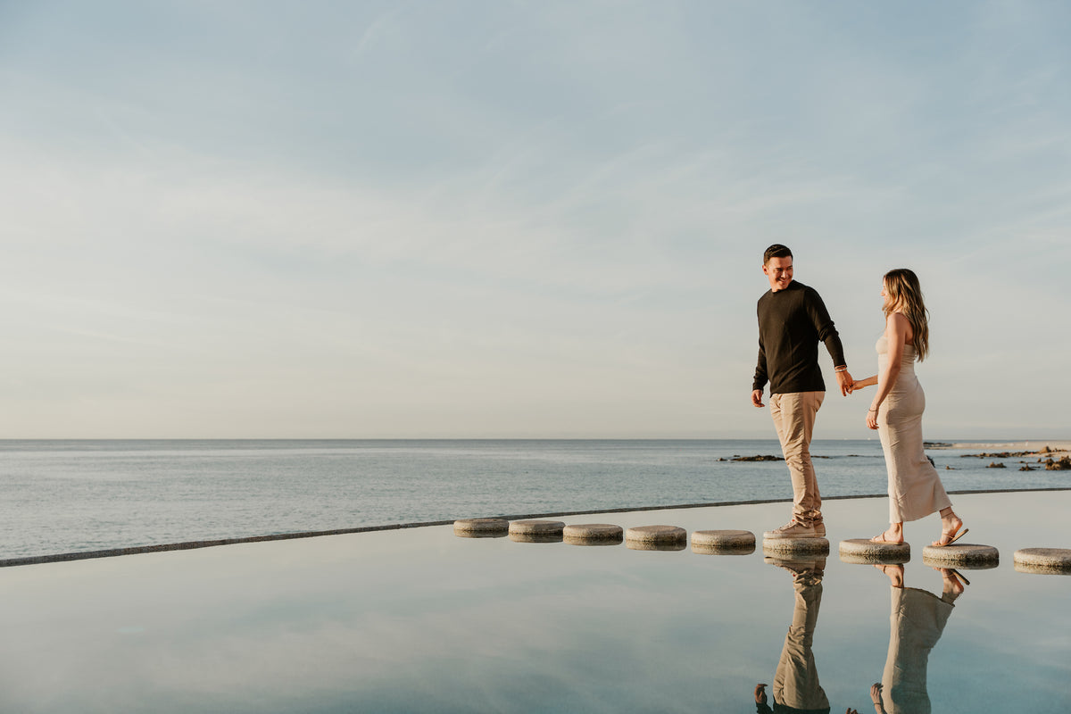 A couple holds hands crossing stepping stones over a reflective pool at Marquis Los Cabos