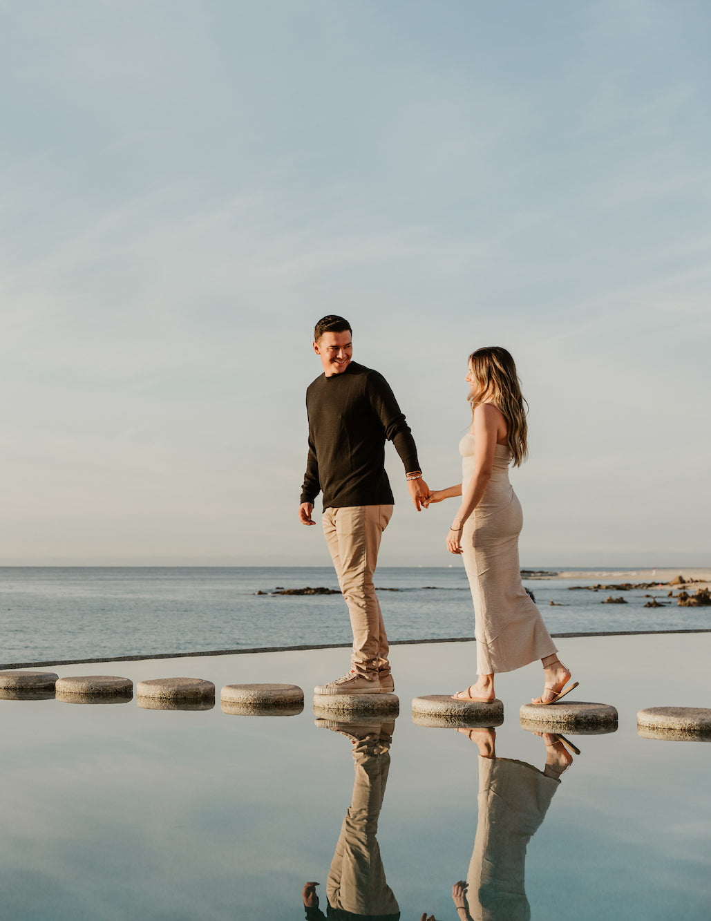 A couple holds hands crossing stepping stones over a reflective pool at Marquis Los Cabos