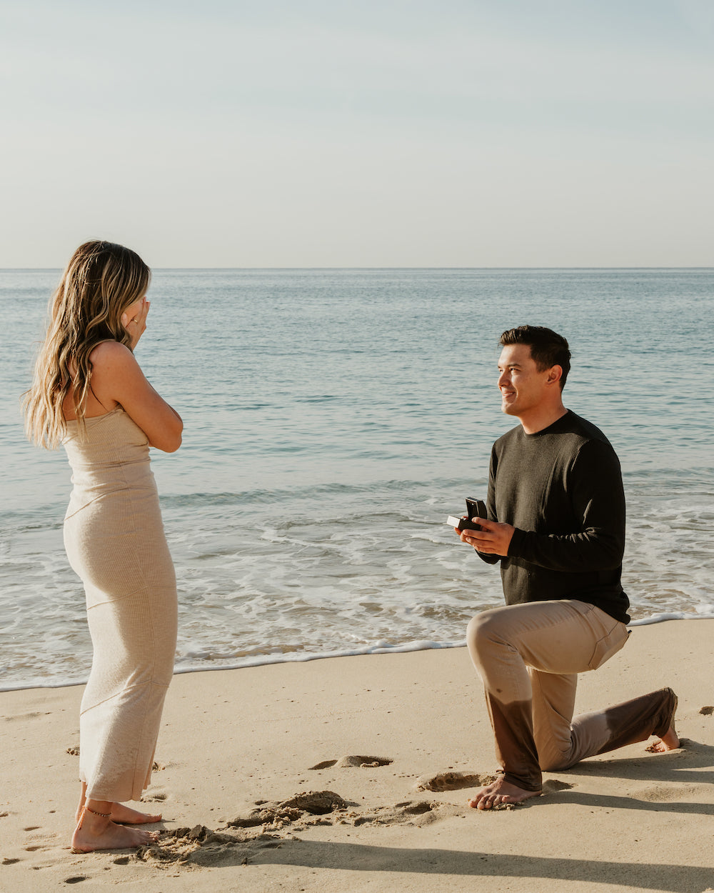 A man kneels on the beach at Marquis Los Cabos, presenting a ring box to a woman