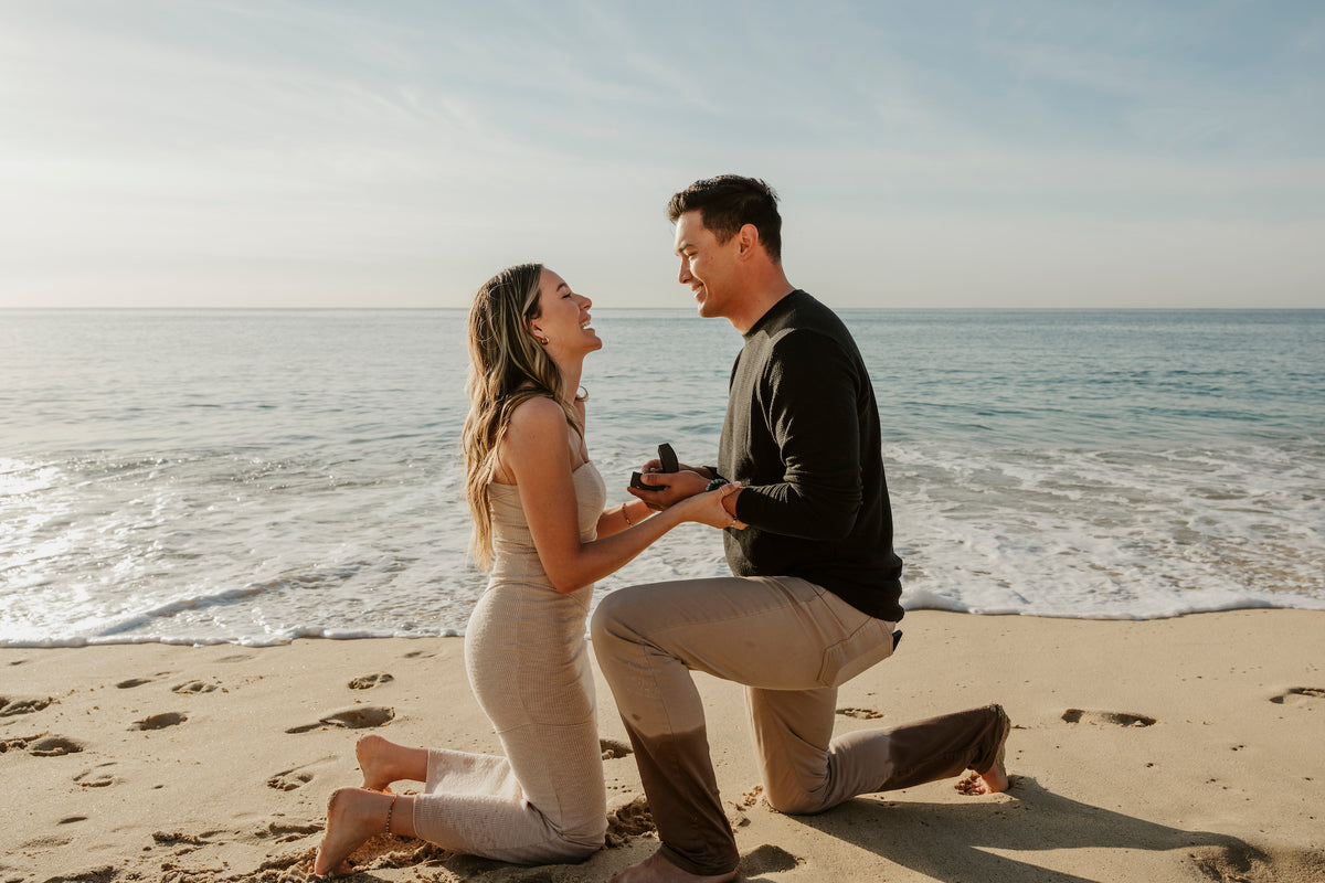 A couple kneels on a beach at Marquis Los Cabos, exchanging a ring box near the ocean