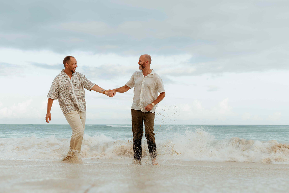 Two men hold hands while walking through ocean waves on the beach