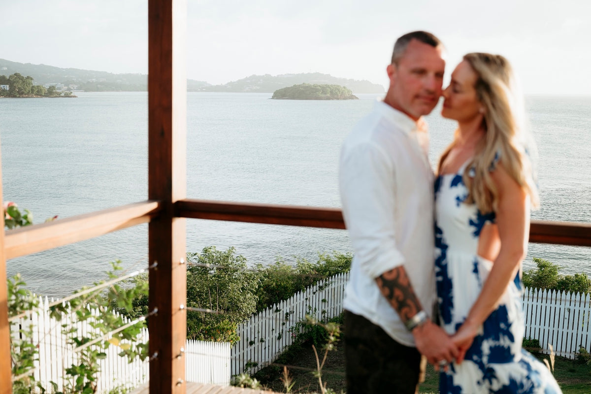 A couple stands close together on a wooden deck overlooking the ocean and coastline