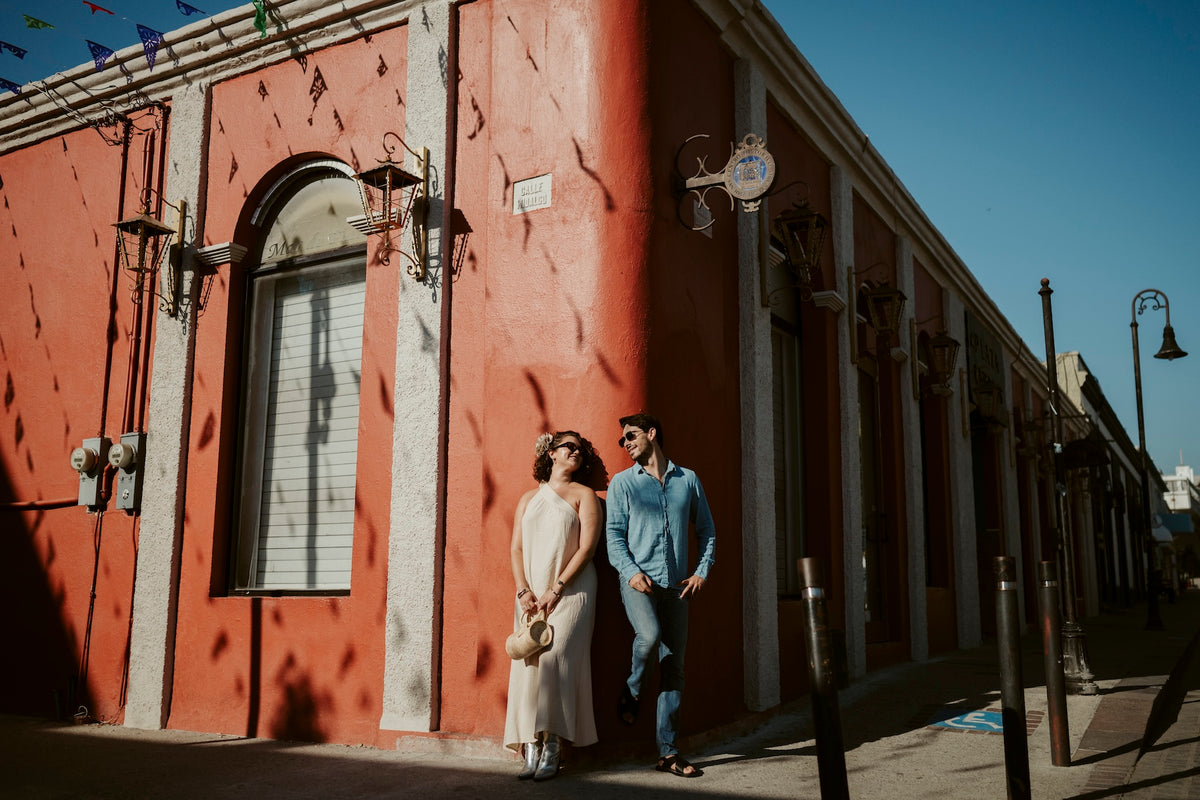 A couple stands against a red building on a sunlit street corner during a DreamArt destination photography session