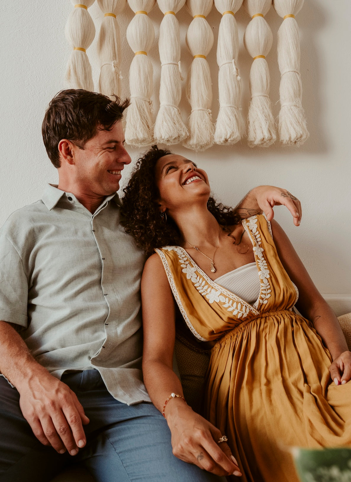 A couple laughing together on a sofa beneath a cream fiber wall hanging