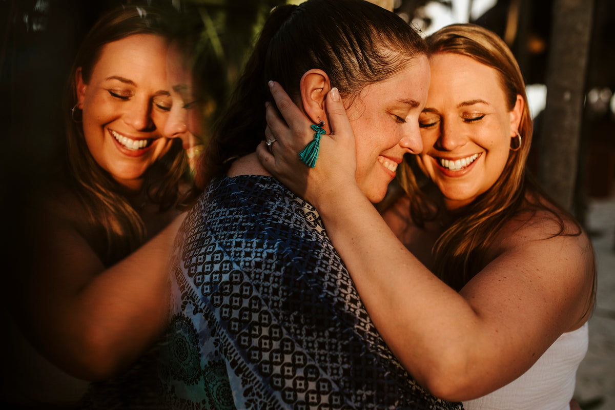 Two women embrace and smile with eyes closed in warm sunlight at Valentin Imperial Riviera Maya, a mirrored reflection visible beside them