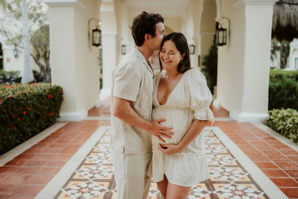 A pregnant woman smiles as a man kisses her head under an archway at Valentin Imperial Riviera Maya
