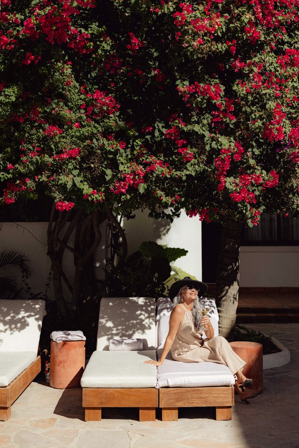A woman in a sun hat sips a martini on a poolside lounge chair beneath bougainvillea at a resort