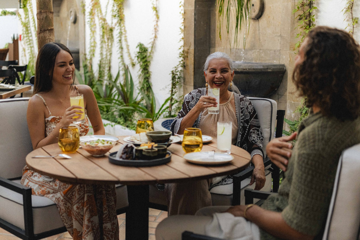 Three women share drinks and food at an outdoor table on a plant-lined patio at DreamArt resort
