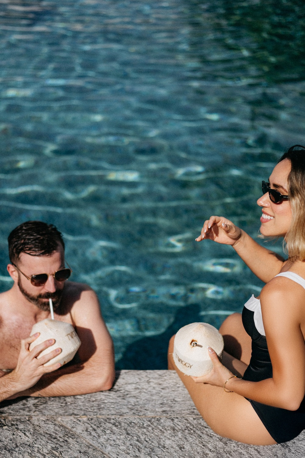 A man and woman relax poolside at a resort, each holding a fresh coconut drink with straws