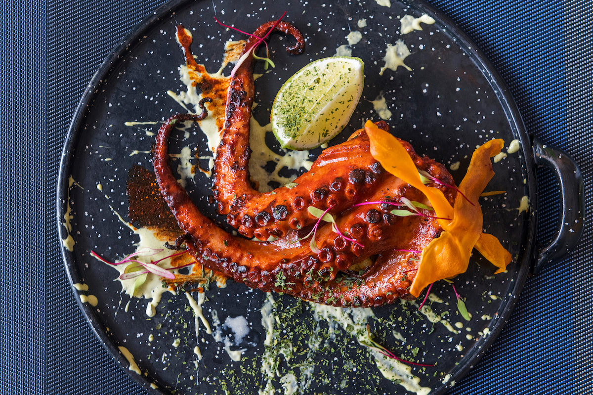 Overhead view of grilled octopus with lime and edible flowers on a black plate, resort food photography