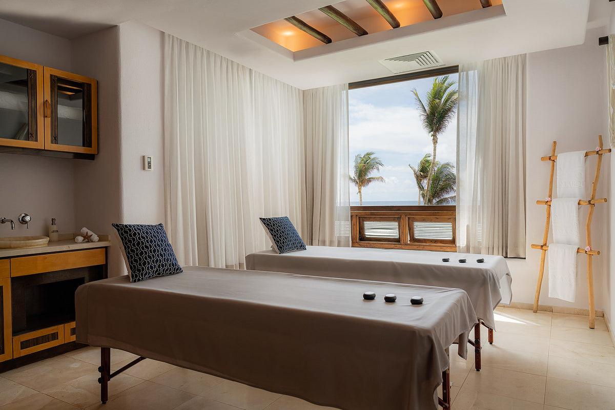 Couples spa treatment room with two massage tables, hot stones and a palm-view window at Marquis Los Cabos