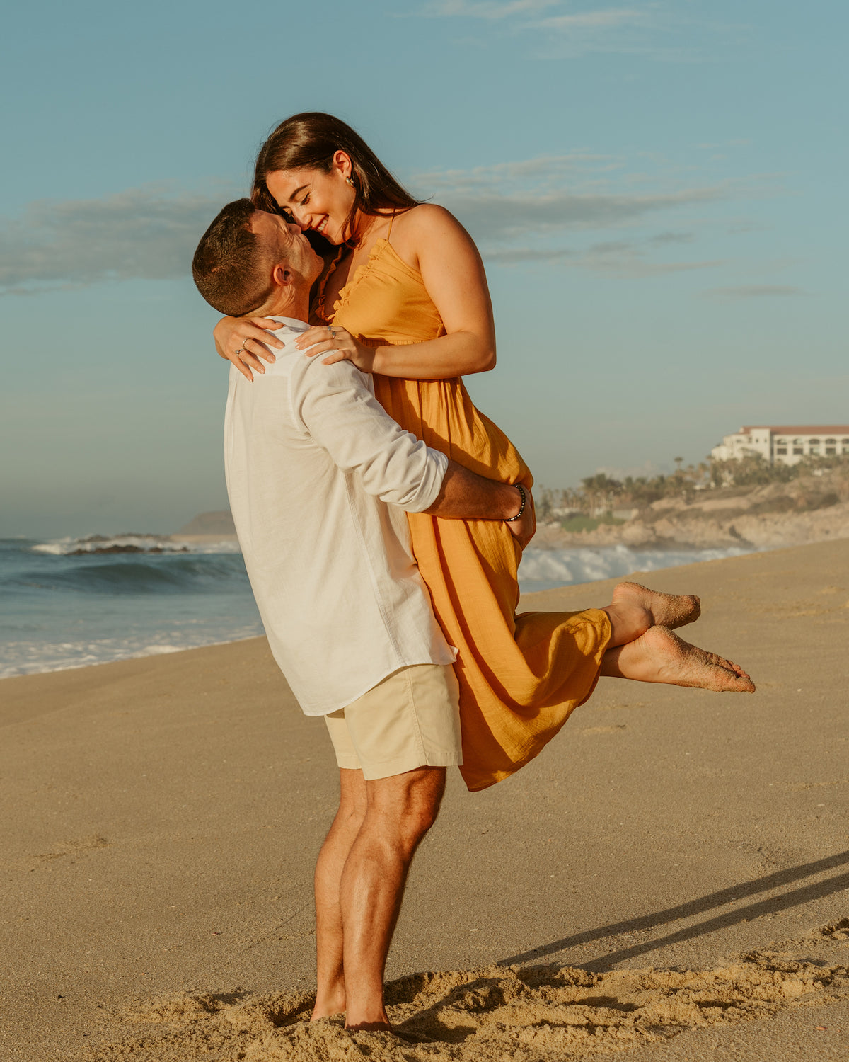 Man lifts his laughing partner in a yellow dress on a sandy beach during a couples photoshoot
