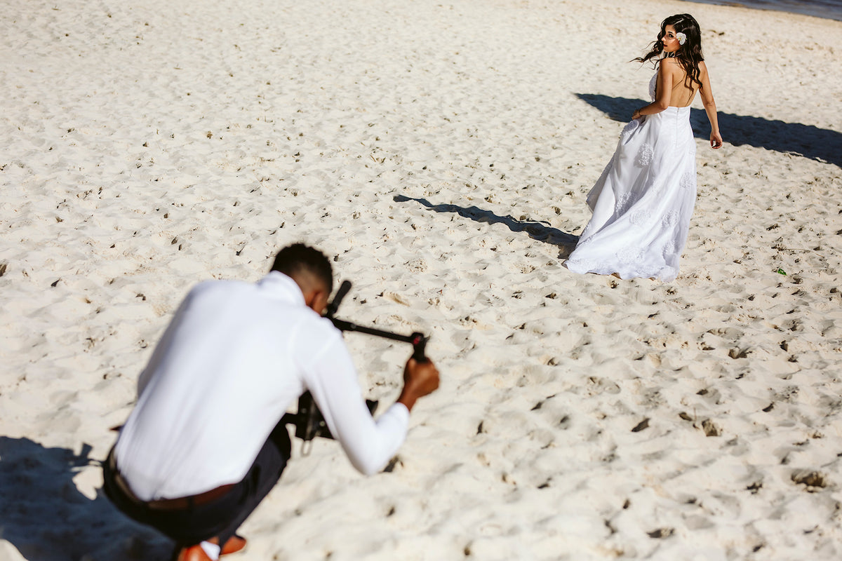 Photographer crouches with a camera rig to film a bride walking across the sand at Moon Palace