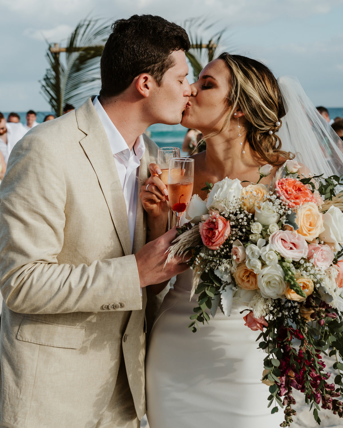 Bride and groom share a kiss holding a peach rose bouquet and champagne on their wedding day