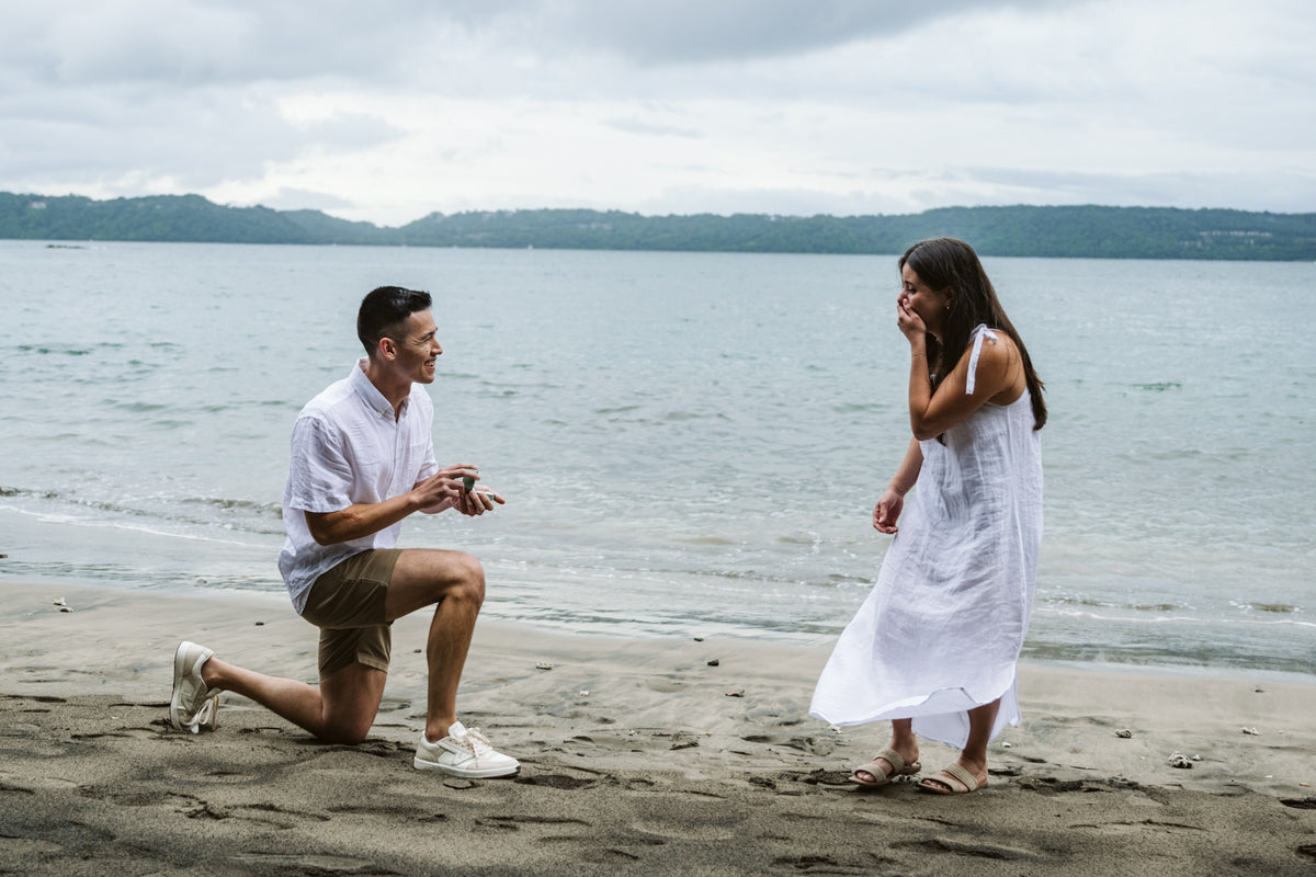Man kneels to propose on a dark sand beach as she covers her face at Secrets Papagayo