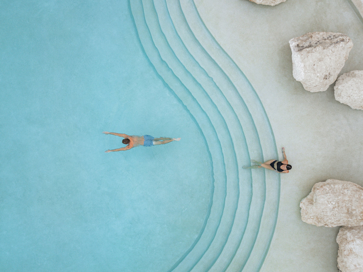 Drone view of a couple swimming in a turquoise pool with curved steps at Secrets Playa Blanca Costa Mujeres