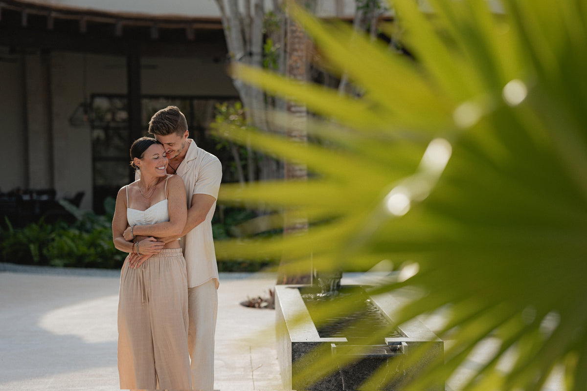 Couple embraces behind palm fronds on the resort plaza during a couples photoshoot at Secrets Playa Blanca Costa Mujeres