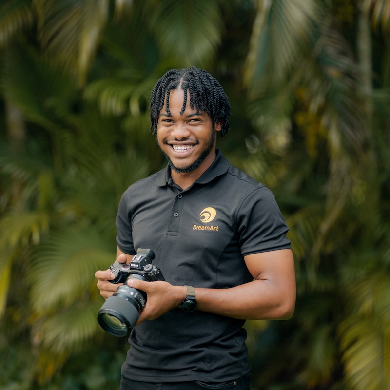 Smiling DreamArt photographer holds his camera in front of lush green palm fronds