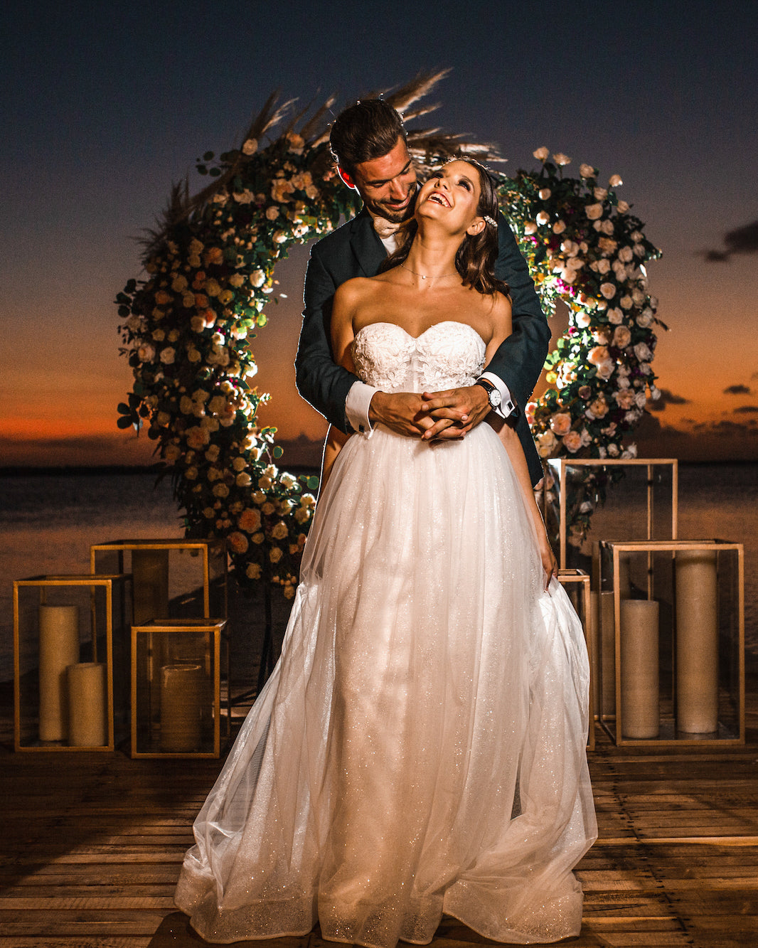 A DreamArt wedding photo of a bride and groom embracing beside a floral arch at sunset