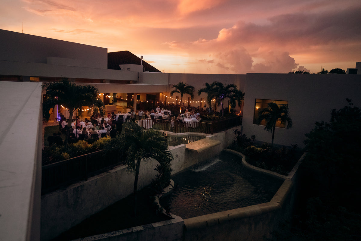Guests dine outdoors at long tables beside a pool with a small waterfall under a pink sunset sky