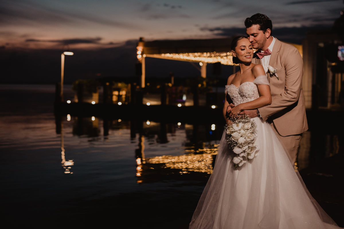 A bride and groom embrace at dusk beside a dock with string lights over calm water