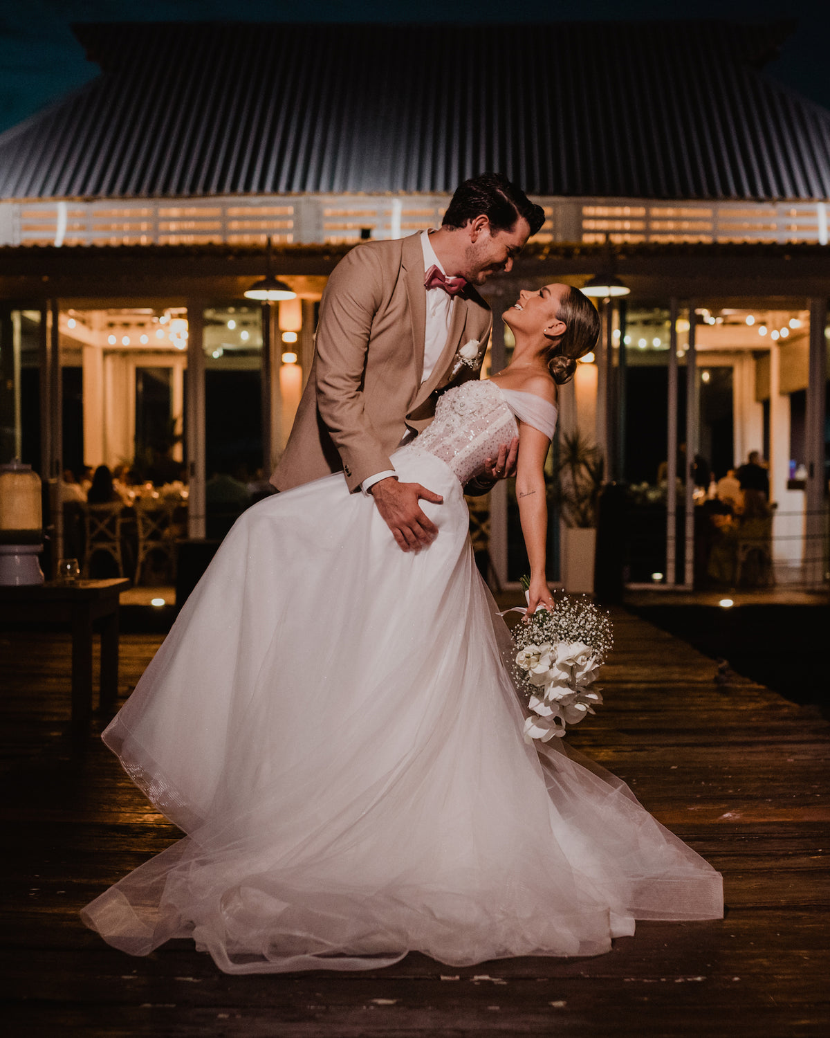 A groom in a tan suit dips a bride on a wooden deck at night, DreamArt wedding photography