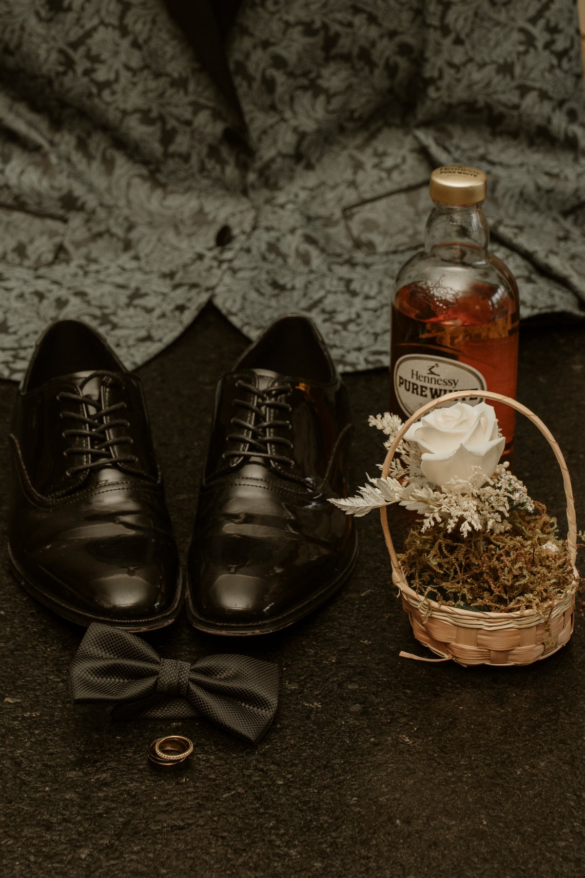 Groom's wedding details with black dress shoes, bow tie, ring, a bottle of whisky and a small flower basket