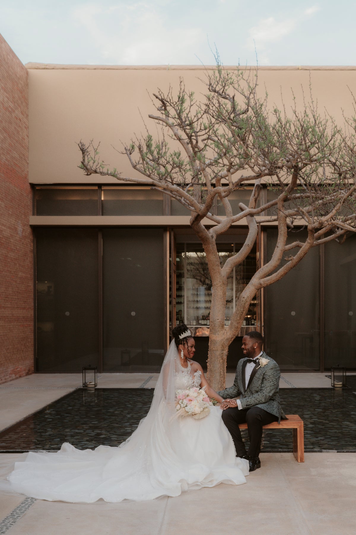 Bride in a ballgown and veil and groom holding hands on a courtyard bench beneath a sculptural bare tree