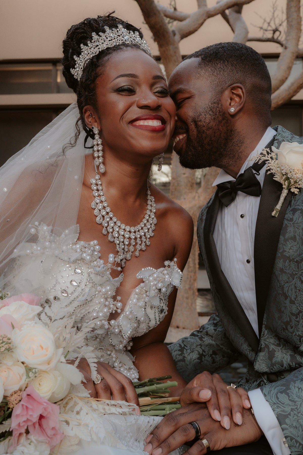 Bride in a beaded gown and tiara and groom in a patterned tuxedo smiling together with a rose bouquet