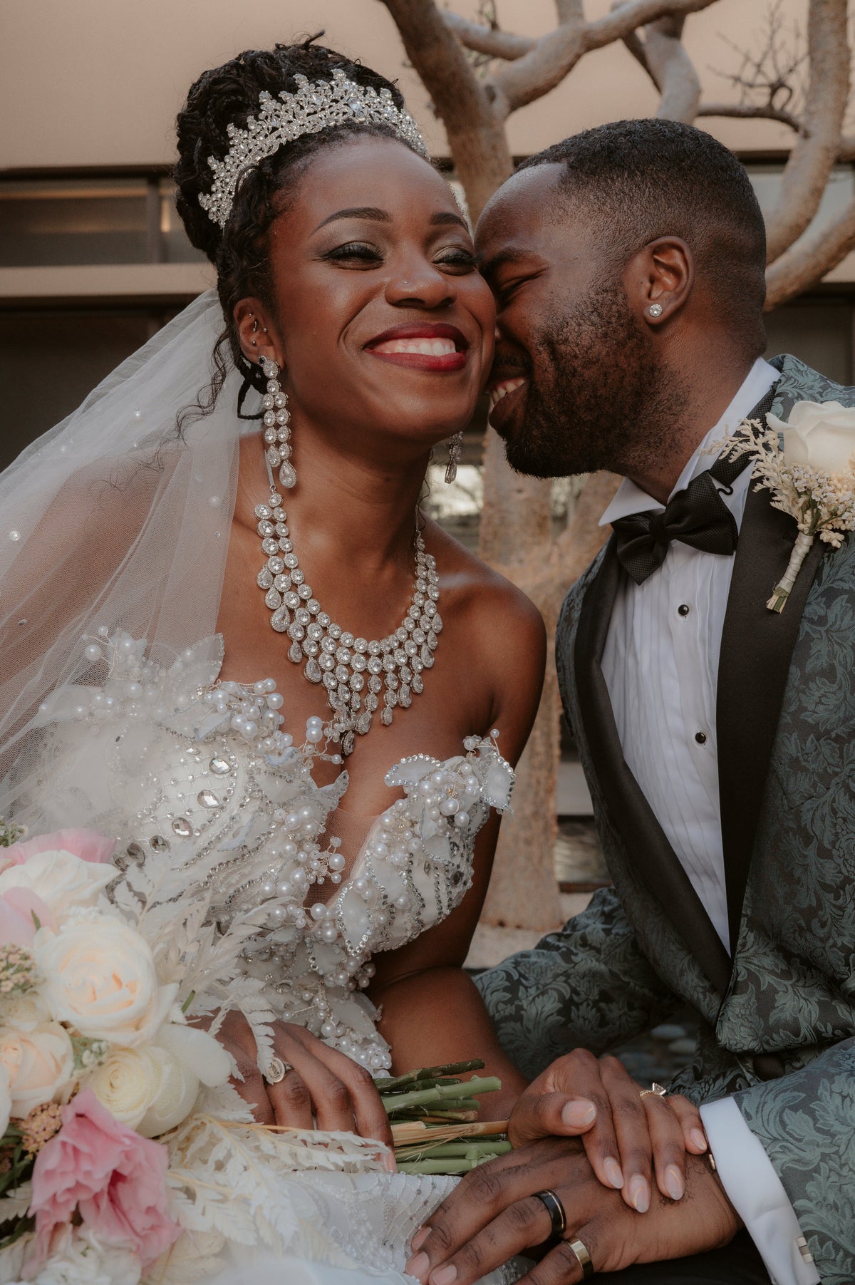 Bride in a beaded gown and crystal tiara and groom in a patterned tuxedo smiling together with a rose bouquet
