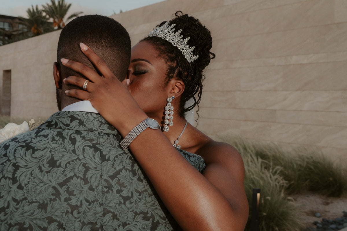 Bride in a crystal tiara embracing a groom in a green damask jacket by a sandstone wall and tall grasses