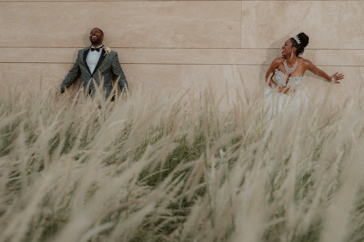 Bride and groom laughing against a sandstone wall behind tall ornamental grasses