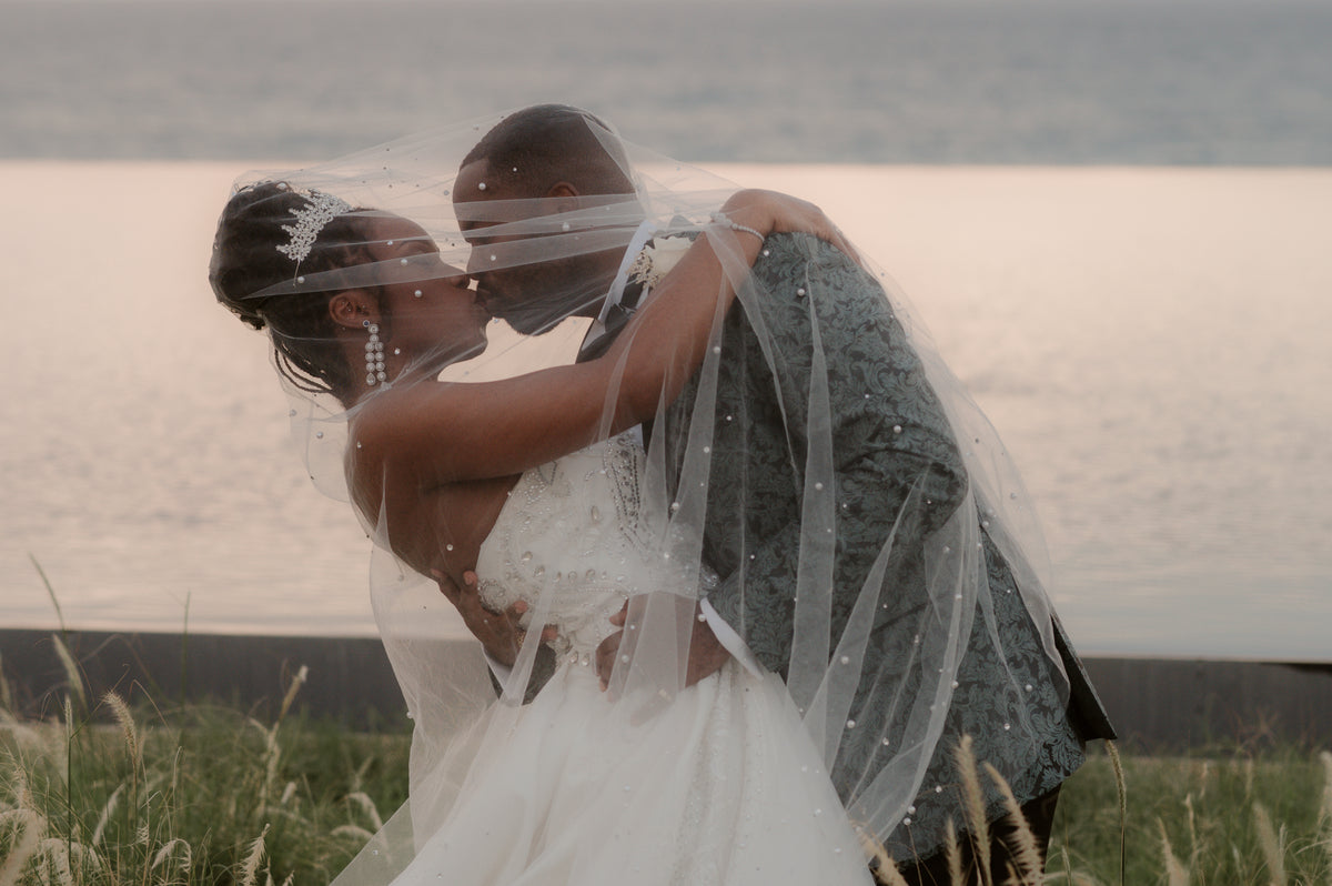 Bride and groom kissing beneath the veil at dusk beside tall grasses and calm water