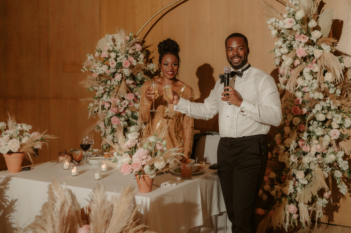 Bride in a gold sequined dress and groom in a bow tie toasting at a reception beside pampas-and-rose arrangements