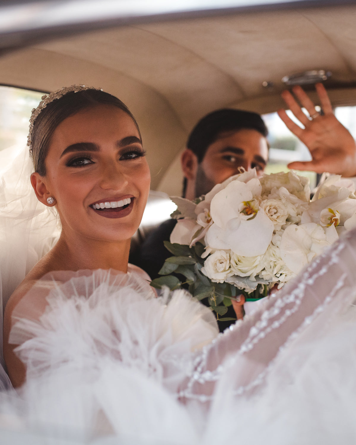 Bride smiling inside a car holding a white bouquet as a groom waves behind her at Grand Fiesta Americana Coral Beach Cancún