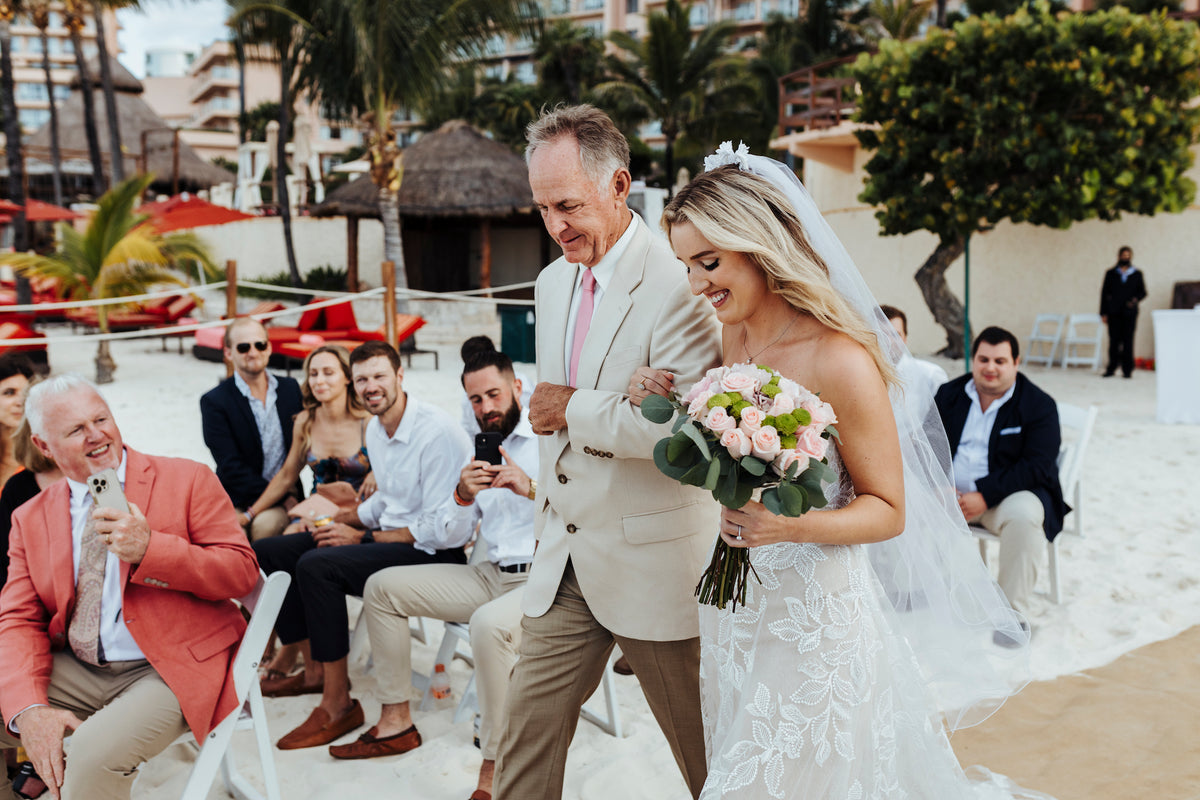 A bride and her father walk down a sandy aisle at Grand Fiesta Americana Coral Beach Cancún as seated guests watch