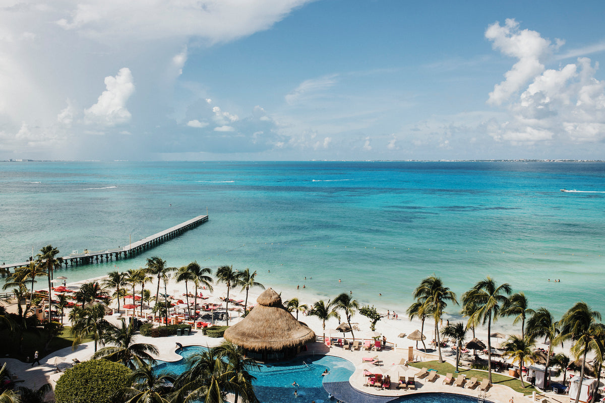 Aerial view of a resort pool, palm trees, and pier along a turquoise Cancún shoreline