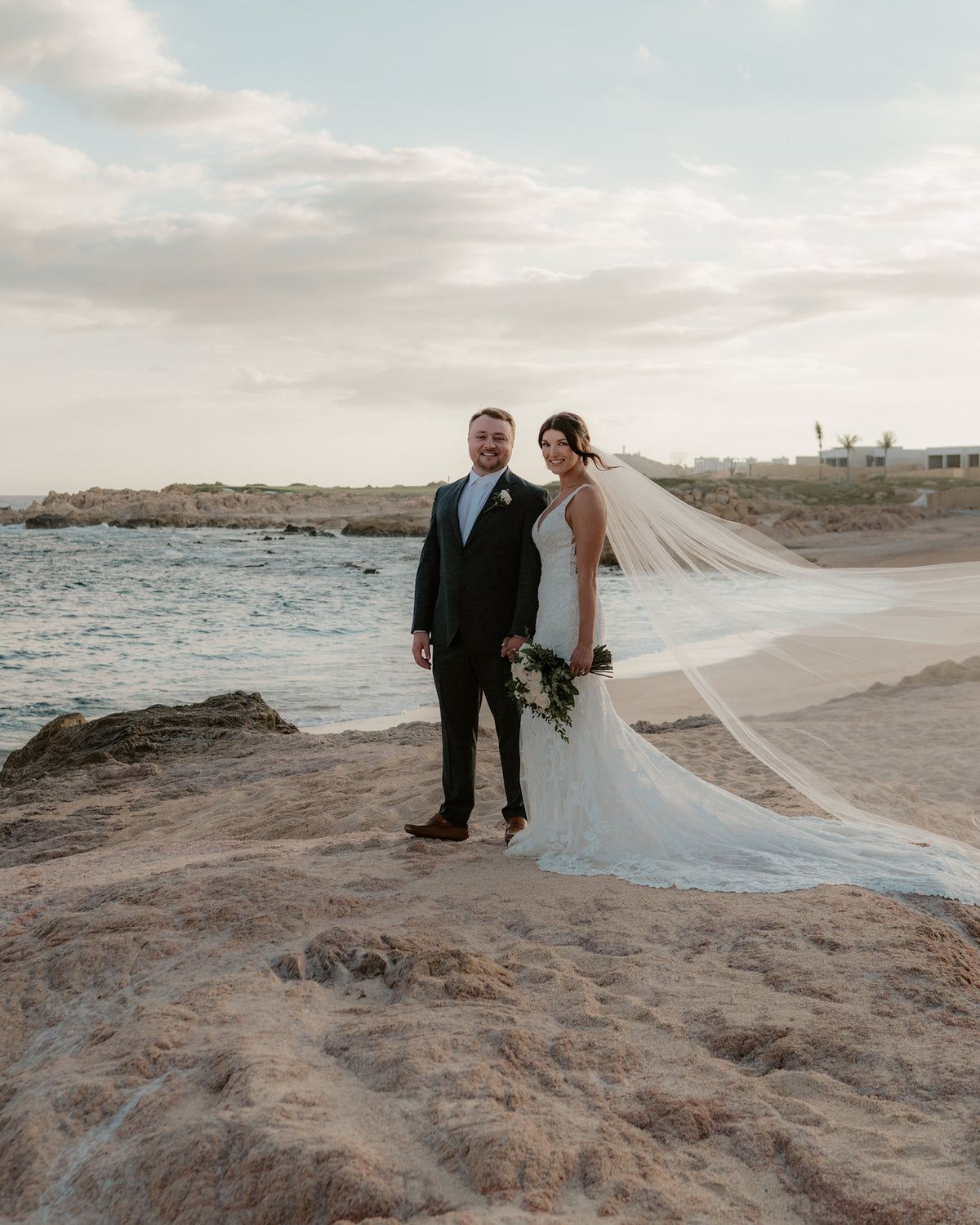 Bride and groom stand on a beach in Los Cabos, her veil blowing in the wind