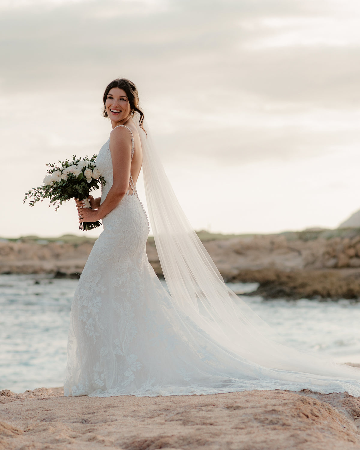 Bride in lace gown and long veil smiling over her shoulder on a beach at Fiesta Americana Los Cabos